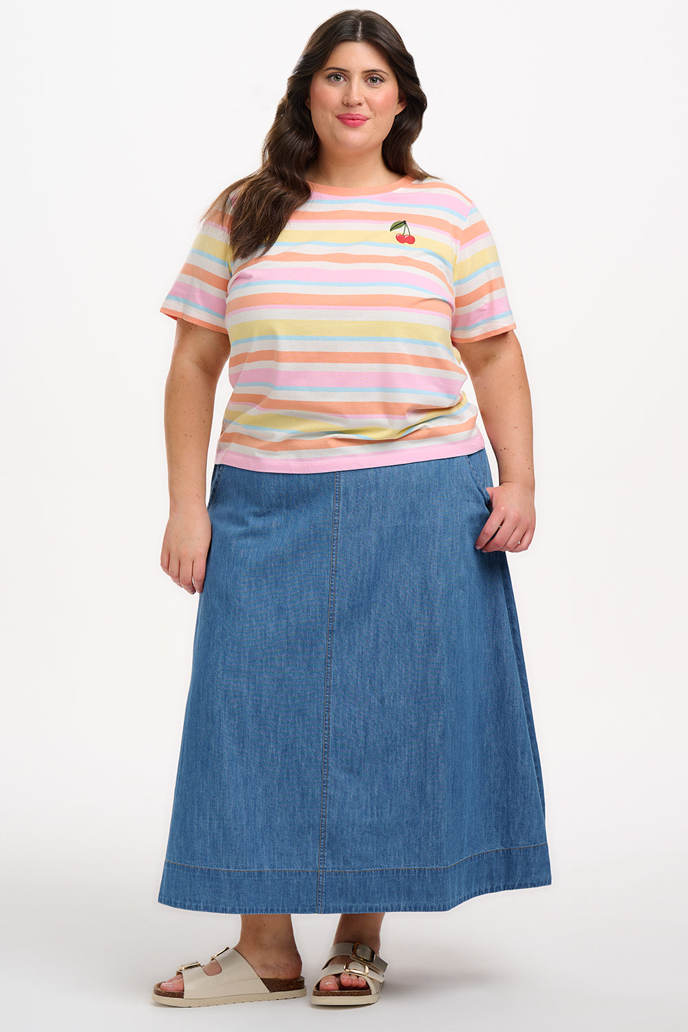 A woman modeling a multicolored striped t-shirt with a small embroidered cherry on the chest, paired with a blue denim skirt.