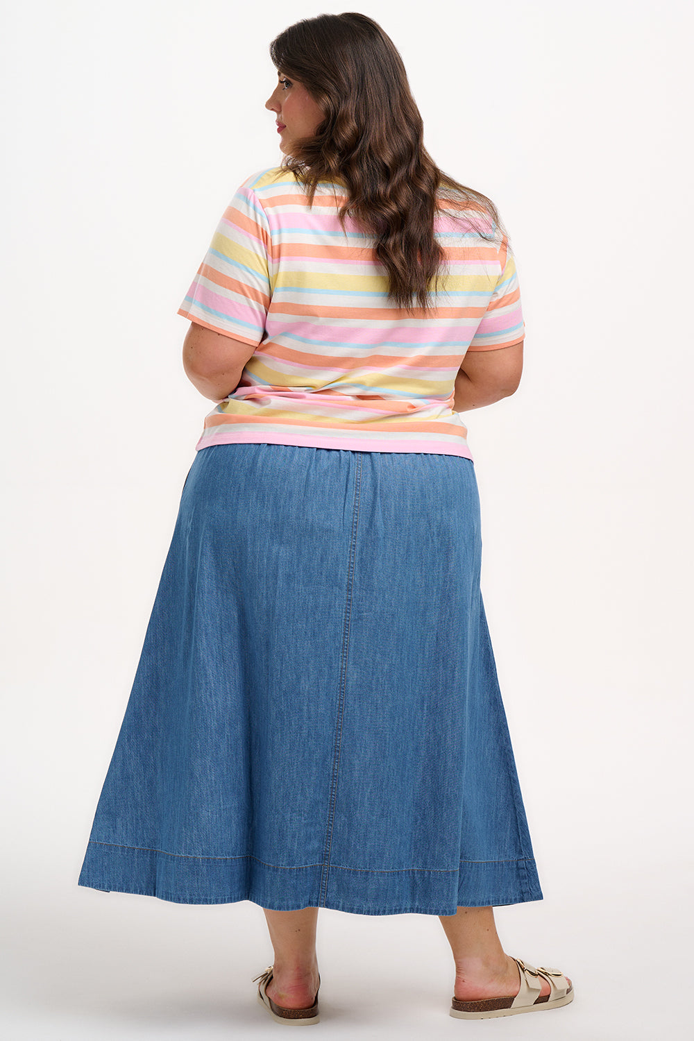A woman modeling a multicolored striped t-shirt with a small embroidered cherry on the chest, paired with a blue denim skirt.