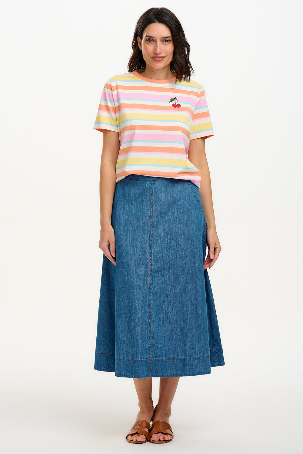 A woman modeling a multicolored striped t-shirt with a small embroidered cherry on the chest, paired with a blue denim skirt.