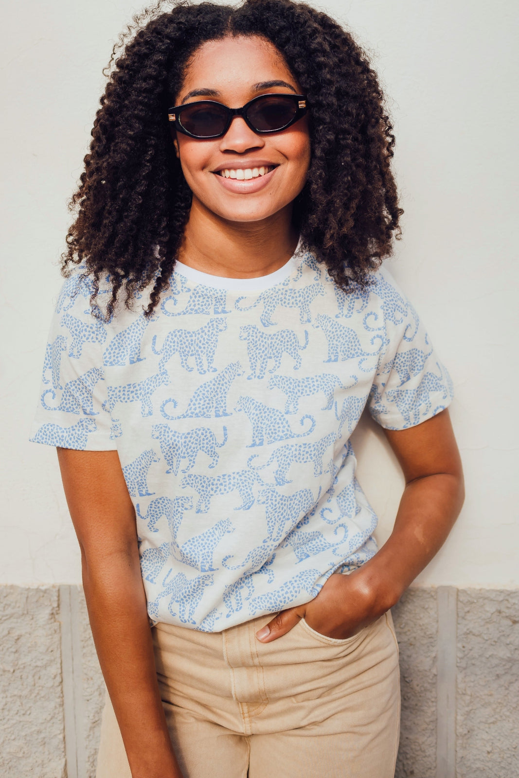 A woman wearing an off-white t-shirt with a blue leopard spot pattern, paired with beige pants and sunglasses, standing against a neutral background.