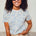 A woman wearing an off-white t-shirt with a blue leopard spot pattern, paired with beige pants and sunglasses, standing against a neutral background.
