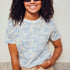 A woman wearing an off-white t-shirt with a blue leopard spot pattern, paired with beige pants and sunglasses, standing against a neutral background.