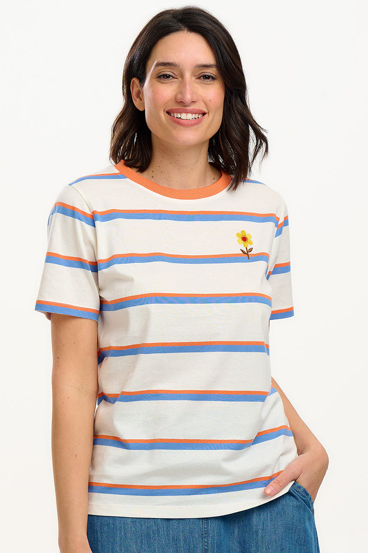 A woman smiling and wearing an off-white t-shirt with blue and orange stripes and a retro style embroidered flower on the chest, paired with a blue skirt.