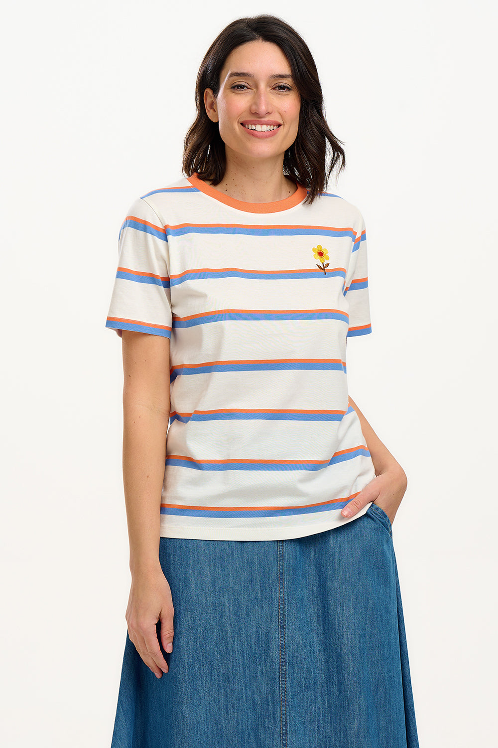 A woman smiling and wearing an off-white t-shirt with blue and orange stripes and a retro style embroidered flower on the chest, paired with a blue skirt.