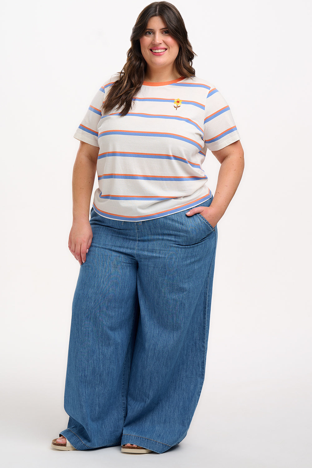 A woman smiling and wearing an off-white t-shirt with blue and orange stripes and a retro style embroidered flower on the chest, paired with a blue skirt.