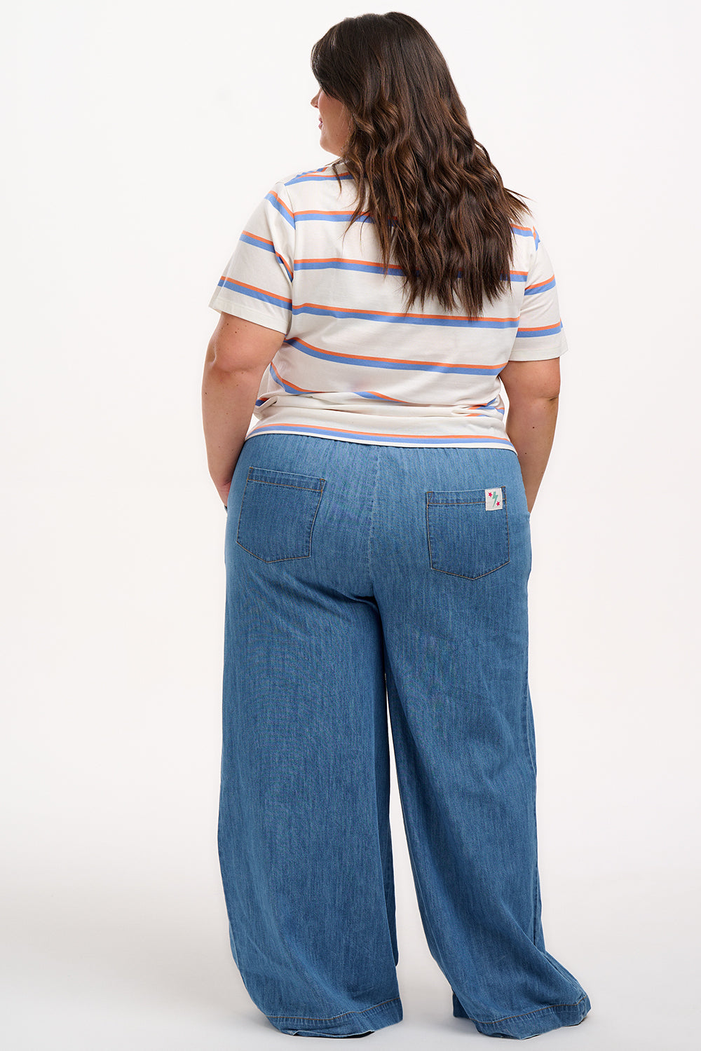 A woman smiling and wearing an off-white t-shirt with blue and orange stripes and a retro style embroidered flower on the chest, paired with a blue skirt.