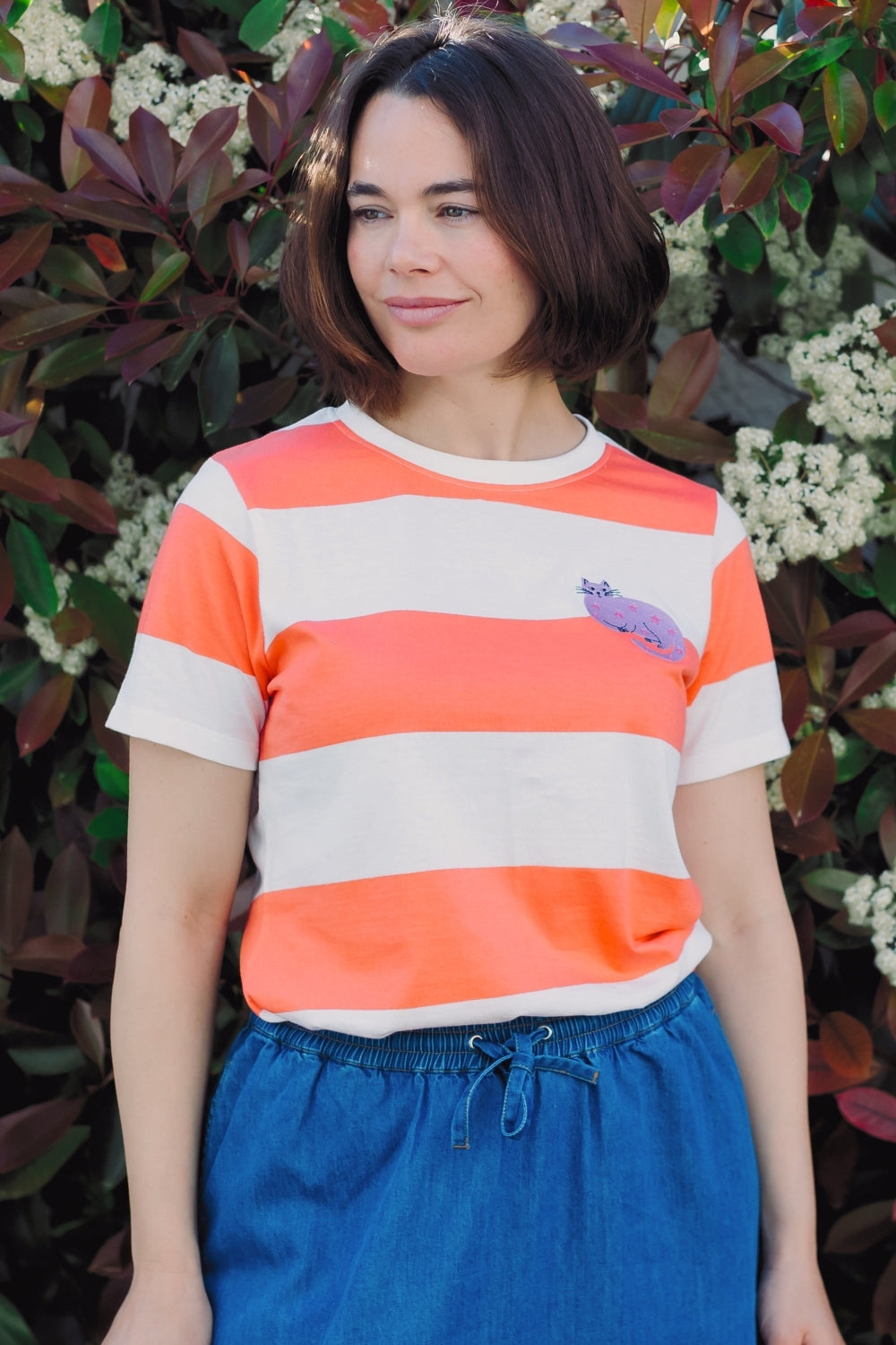 Person wearing an orange and white striped shirt with a purple logo, standing in front of greenery.