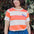 Person wearing an orange and white striped shirt with a purple logo, standing in front of greenery.