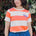 Person wearing an orange and white striped shirt with a purple logo, standing in front of greenery.