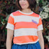 Person wearing an orange and white striped shirt with a purple logo, standing in front of greenery.