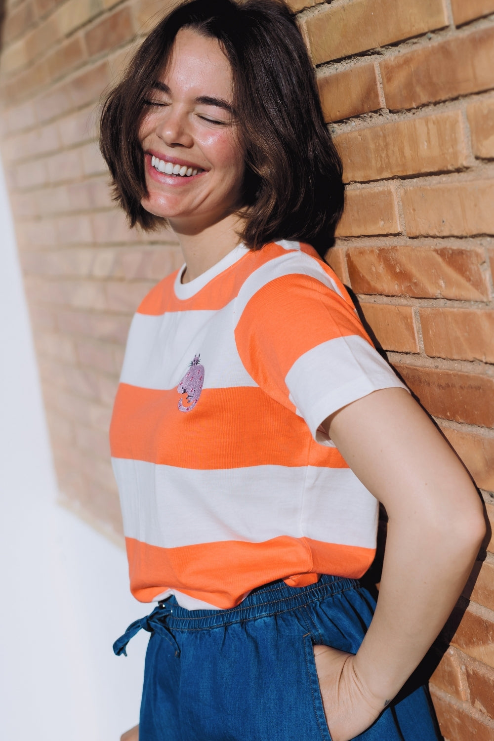 Woman wearing an orange and white striped shirt with a graphic design, standing against a brick wall.