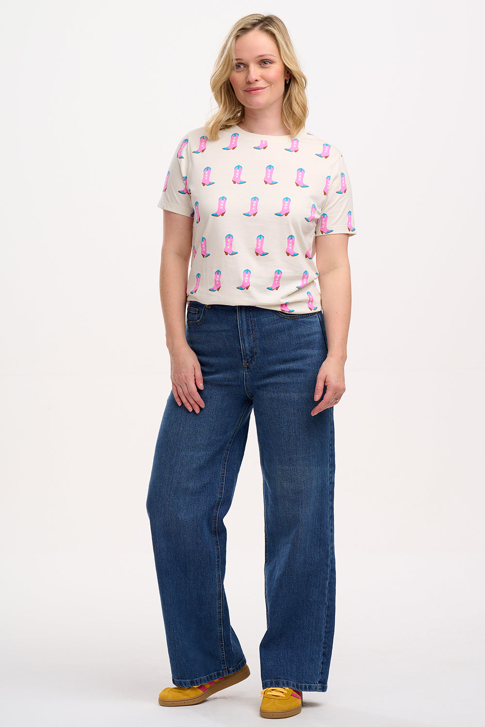 A woman smiling and wearing an off-white t-shirt with a pattern of cowgirl boots in pink and blue, paired with jeans.