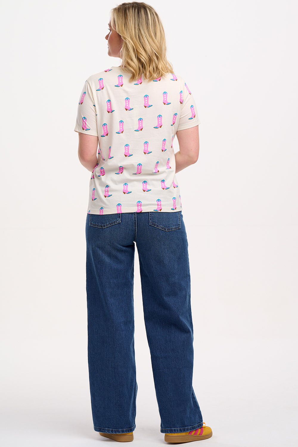A woman smiling and wearing an off-white t-shirt with a pattern of cowgirl boots in pink and blue, paired with jeans.