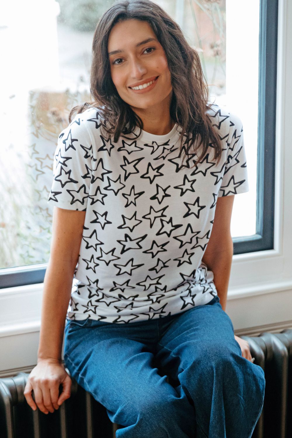 Woman wearing a white t-shirt with black star patterns sitting by a window.