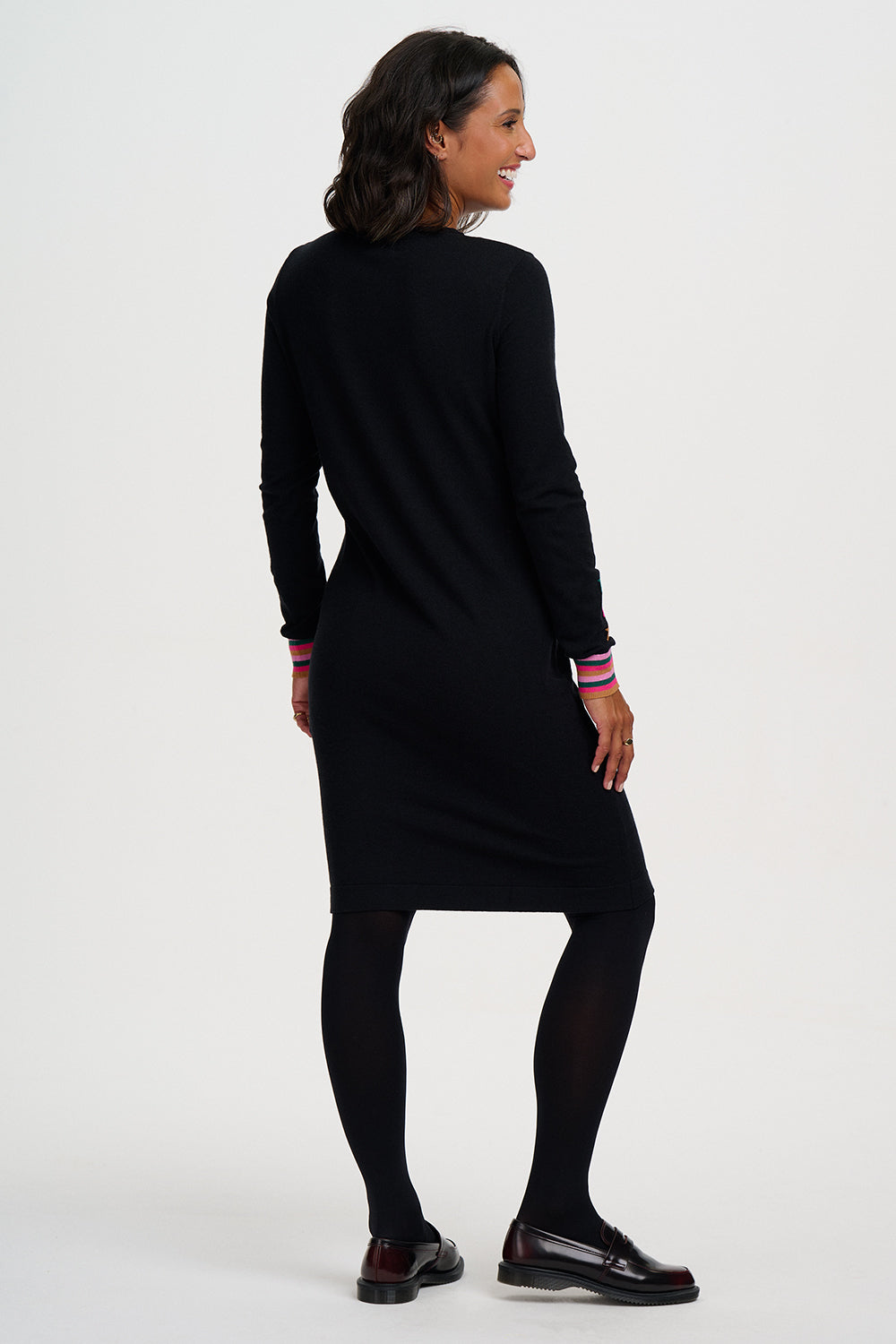 A woman modeling a black long-sleeve knit dress with striped cuffs, paired with black tights and black loafers, shown from a side-back angle against a plain white studio background.
