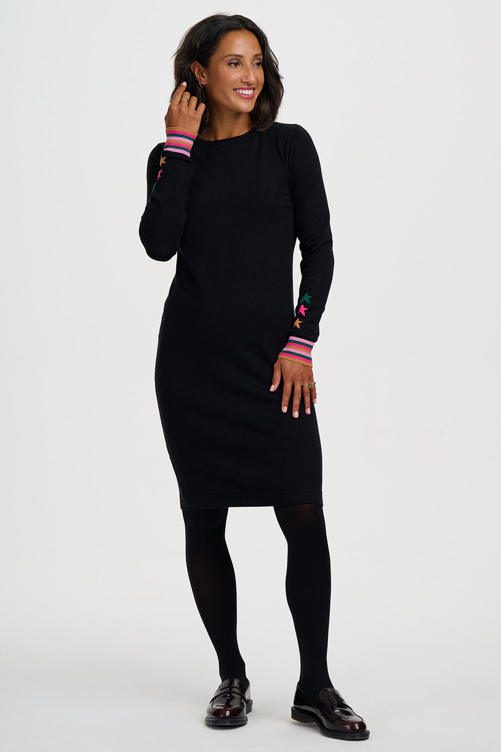 A woman modeling a black long-sleeve knit dress with colorful striped cuffs and embroidered sleeve details, paired with black tights and black shoes, against a plain light gray studio background.