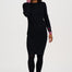 A woman modeling a black long-sleeve knit dress with pink and white striped cuffs, paired with black tights and black shoes, against a plain white studio background.
