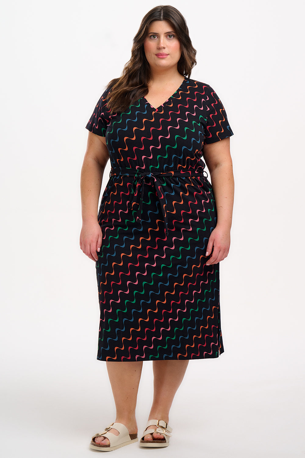 A model wearing a midi-length jersey dress with a V-neck, tie belt, and undulating multicoloured wave print. The dress is primarily black with red, pink, green, blue, and orange patterns.
