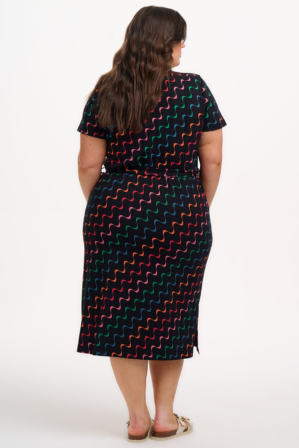 A model wearing a midi-length jersey dress with a V-neck, tie belt, and undulating multicoloured wave print. The dress is primarily black with red, pink, green, blue, and orange patterns.
