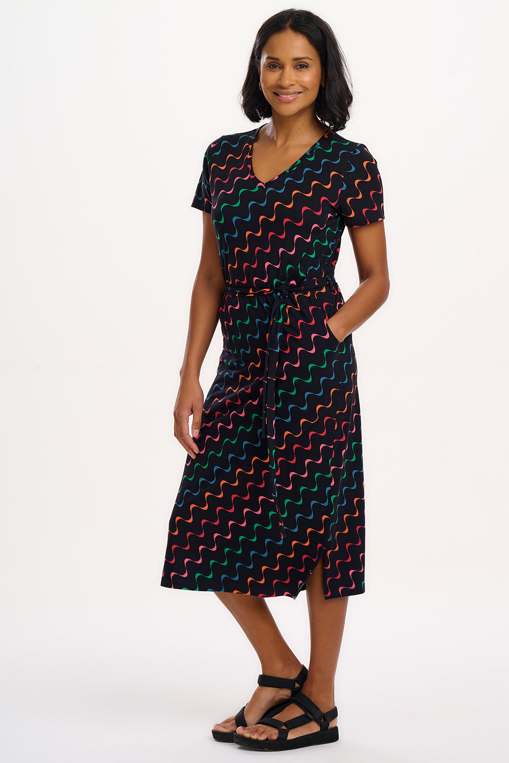 A model wearing a midi-length jersey dress with a V-neck, tie belt, and undulating multicoloured wave print. The dress is primarily black with red, pink, green, blue, and orange patterns.