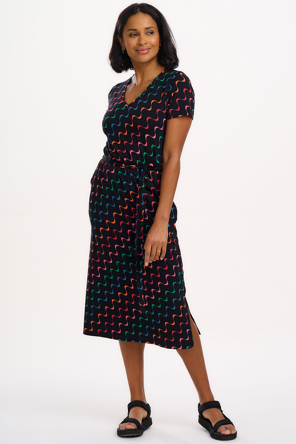 A model wearing a midi-length jersey dress with a V-neck, tie belt, and undulating multicoloured wave print. The dress is primarily black with red, pink, green, blue, and orange patterns.