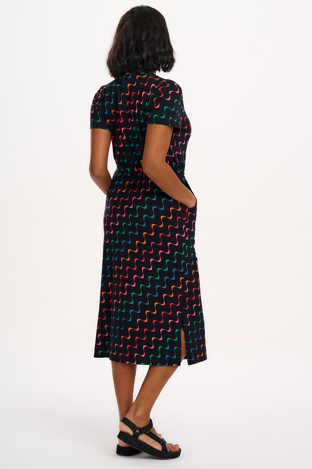 A model wearing a midi-length jersey dress with a V-neck, tie belt, and undulating multicoloured wave print. The dress is primarily black with red, pink, green, blue, and orange patterns.