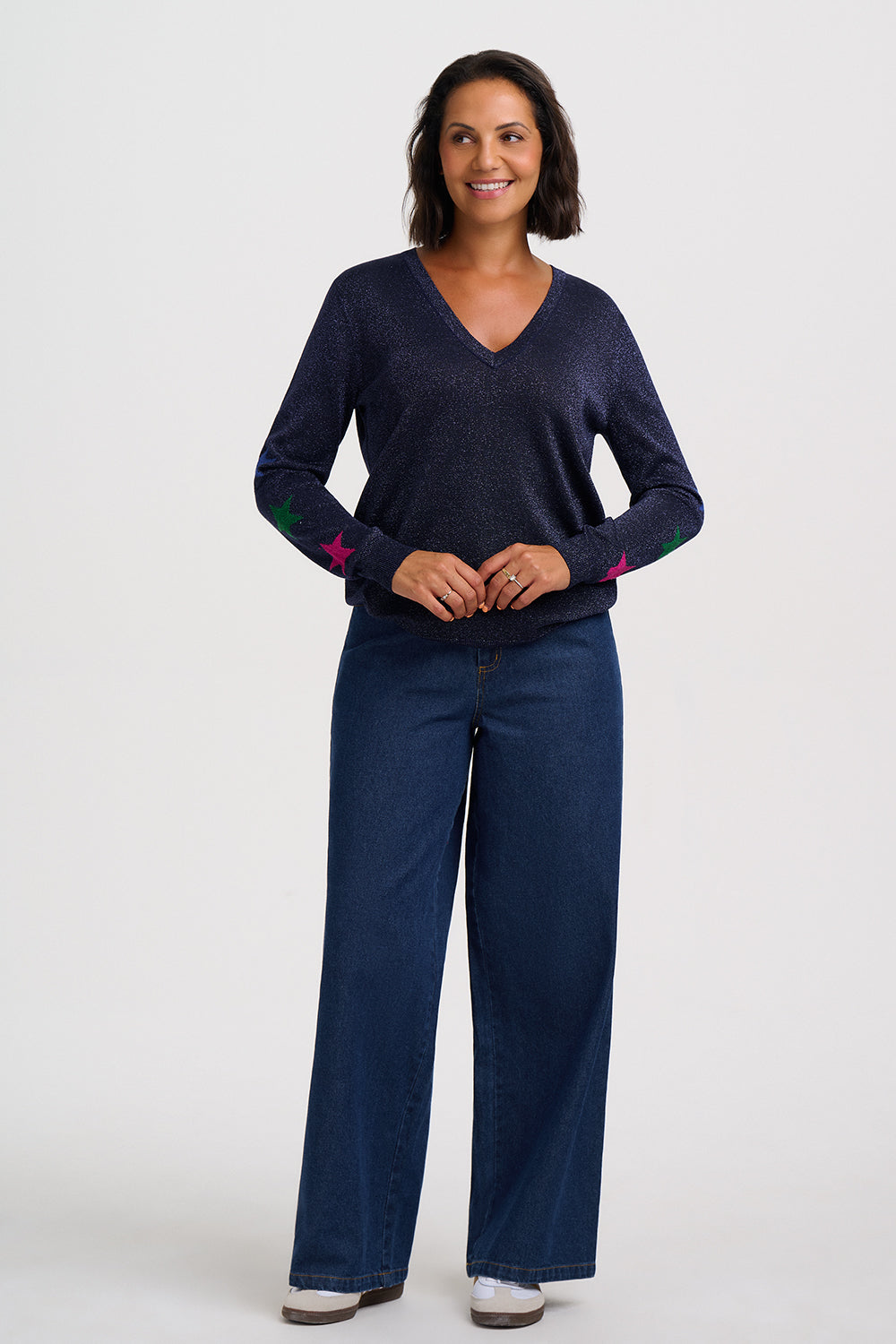 A woman modeling a dark navy blue knit V-neck sweater with colorful embroidered star details on the sleeves, paired with dark blue jeans, against a plain light gray studio background.