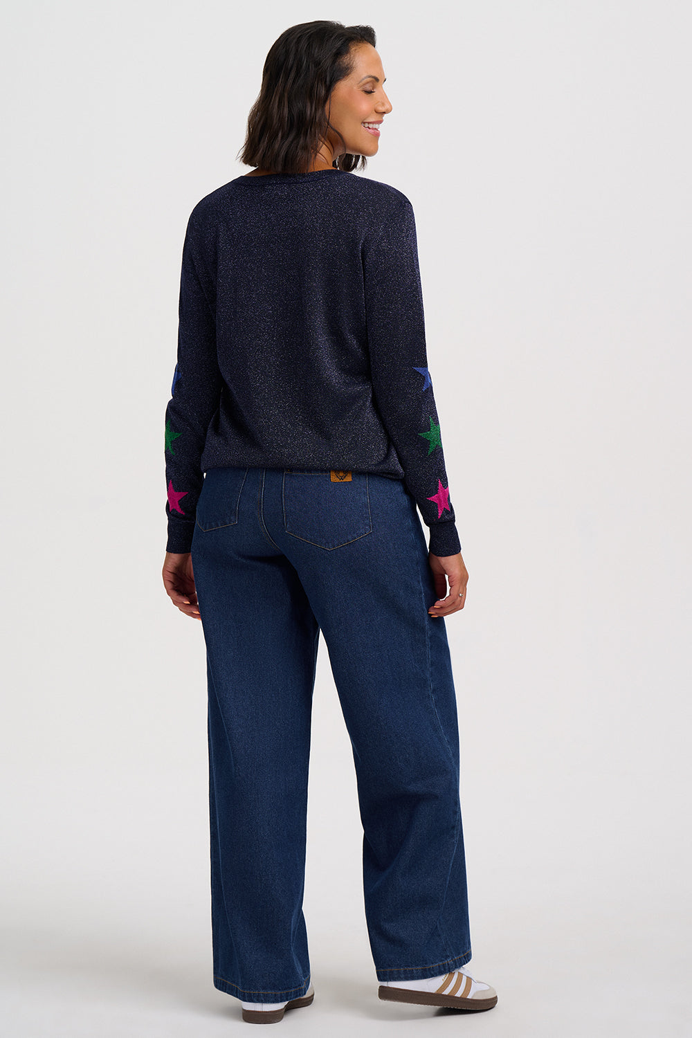 A woman shown from the back wearing a dark navy knit sweater with colorful embroidered star details on the sleeves and dark blue wide-leg jeans, standing against a light gray studio background.
