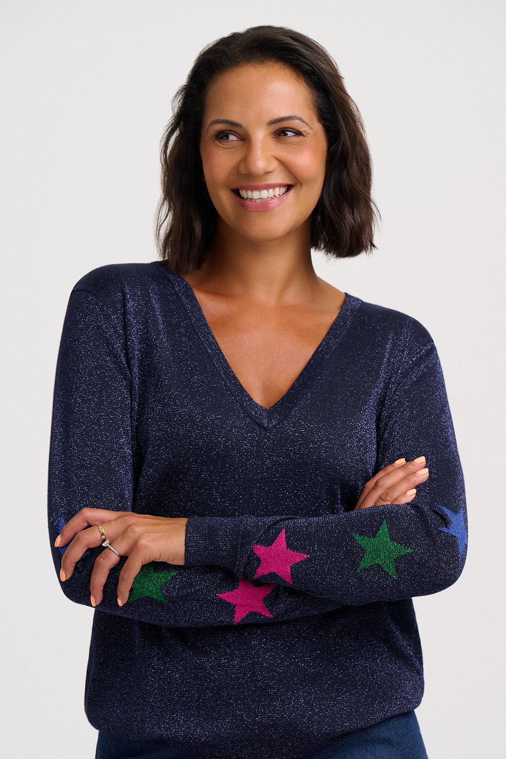A smiling woman with short dark hair wearing a navy blue V-neck sweater with colorful star patches on the sleeves, posed against a light gray studio background.