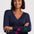A smiling woman with short dark hair wearing a navy blue V-neck sweater with colorful star patches on the sleeves, posed against a light gray studio background.