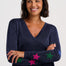 A smiling woman with short dark hair wearing a navy blue V-neck sweater with colorful star patches on the sleeves, posed against a light gray studio background.