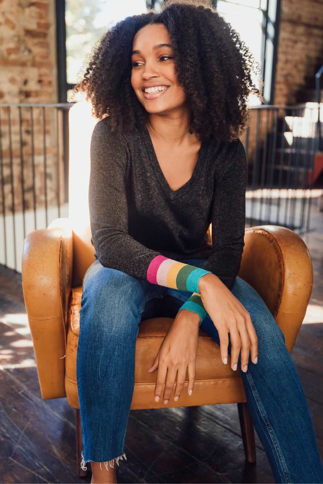 Woman sitting on a brown leather chair wearing a dark gray sweater and blue jeans with colorful arm sleeves.