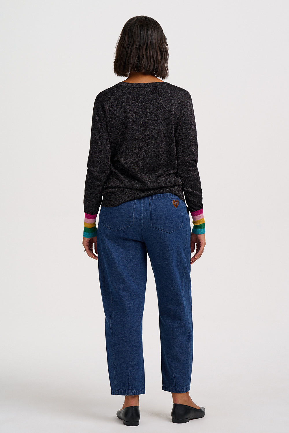 A model shown from the back wearing a dark charcoal knit long-sleeve top with colorful striped cuffs, paired with blue jeans and black flats, against a plain white studio background.