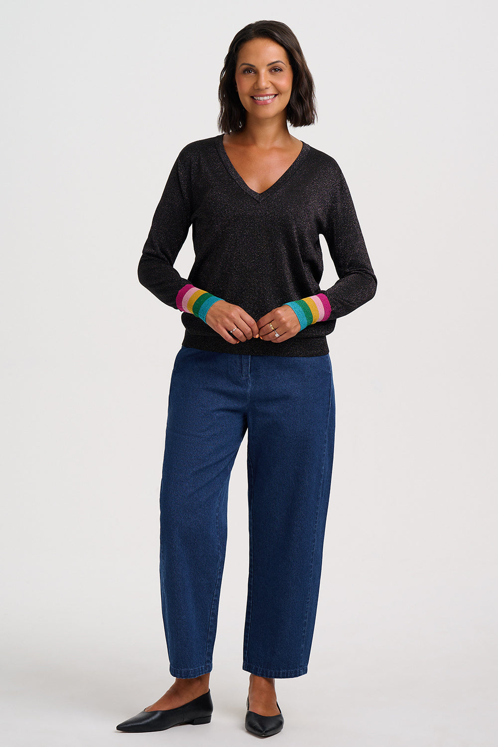 A woman modeling a dark charcoal gray V-neck knit sweater with vibrant rainbow-striped cuffs, paired with dark blue jeans and black flats, against a light gray studio background.