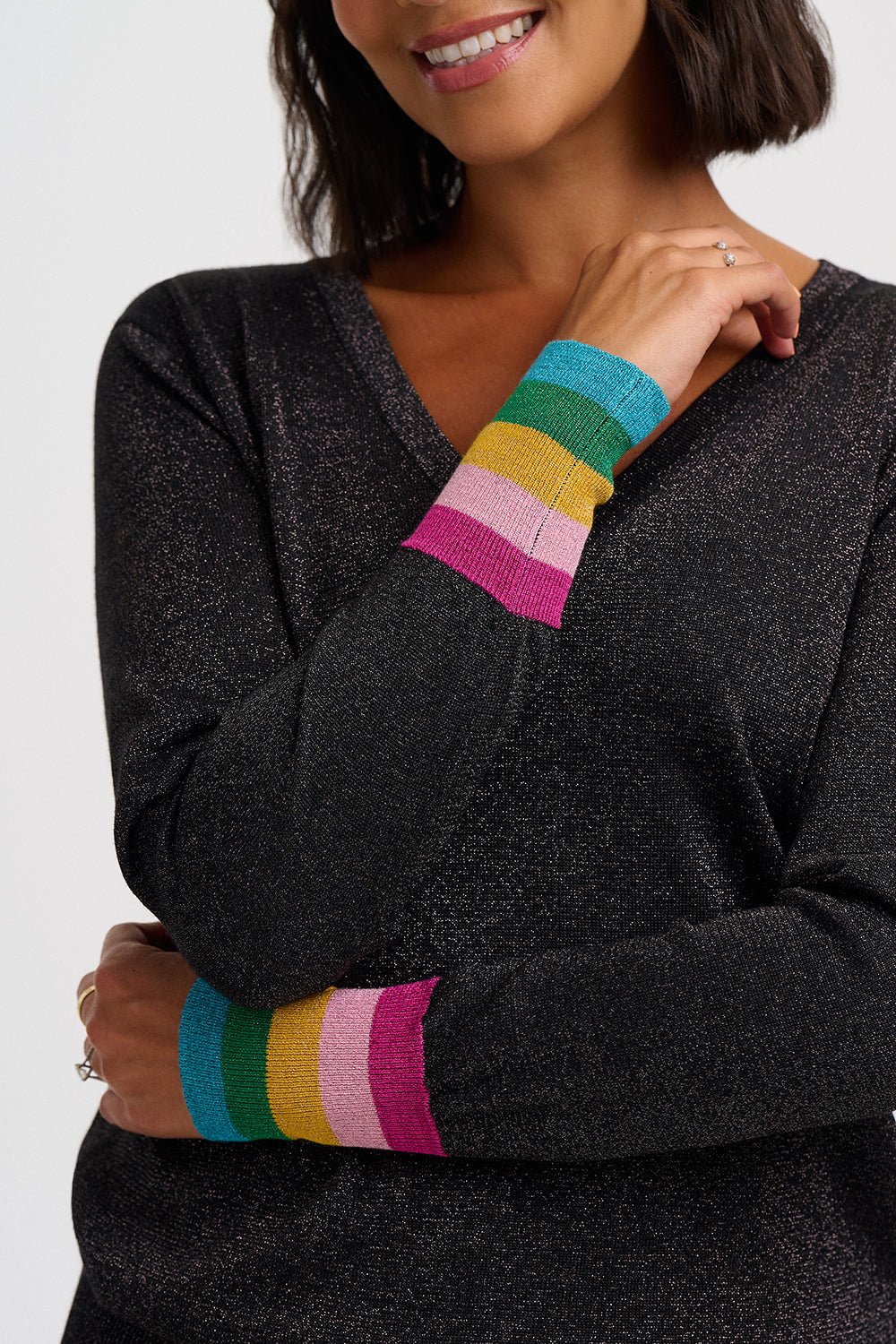 A woman wearing a dark charcoal gray knit sweater with vibrant rainbow-striped ribbed cuffs in teal, green, yellow, pink, and white, shown against a light gray studio background.