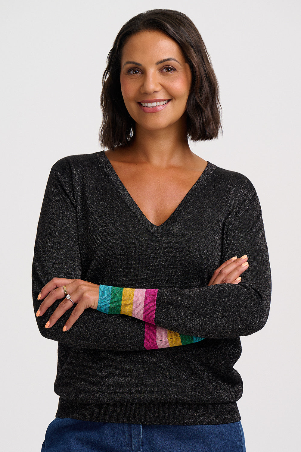 A woman with short dark hair smiling, wearing a dark charcoal gray V-neck sweater with colorful striped knit cuffs in green, yellow, orange, pink, and blue, paired with blue jeans against a light gray background.