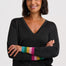 A woman with short dark hair smiling, wearing a dark charcoal gray V-neck sweater with colorful striped knit cuffs in green, yellow, orange, pink, and blue, paired with blue jeans against a light gray background.