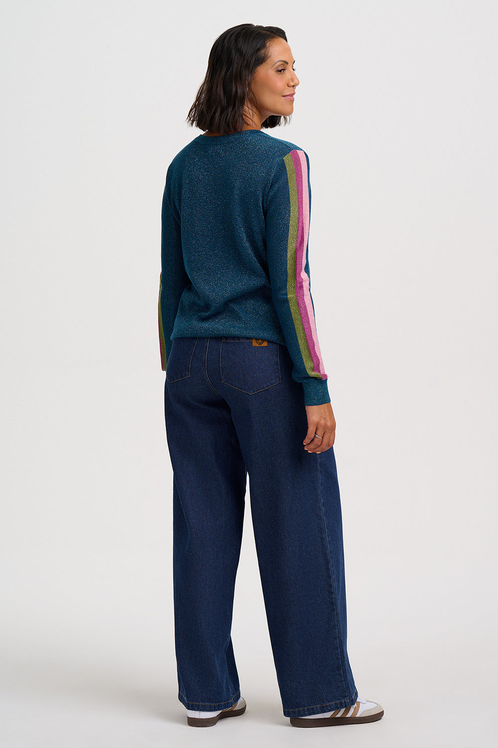 A woman shown from the back wearing a dark teal knit sweater with colorful vertical stripes down the sleeves and dark blue wide-leg jeans, photographed against a plain light gray studio background.