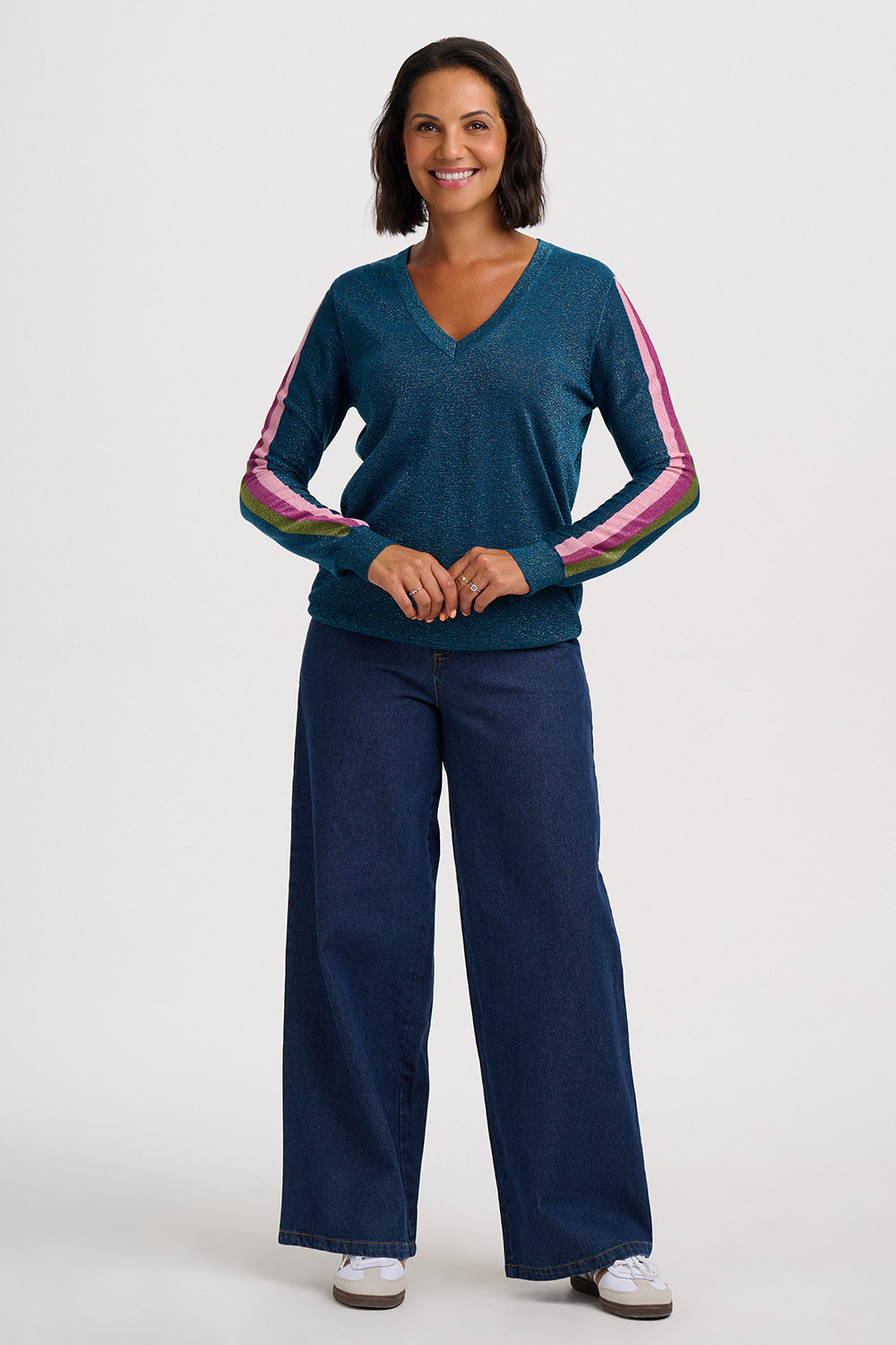 A woman modeling a teal blue long-sleeve knit sweater with pink and green striped accents down the sleeves, paired with dark blue wide-leg jeans and white sneakers, against a light gray studio background.