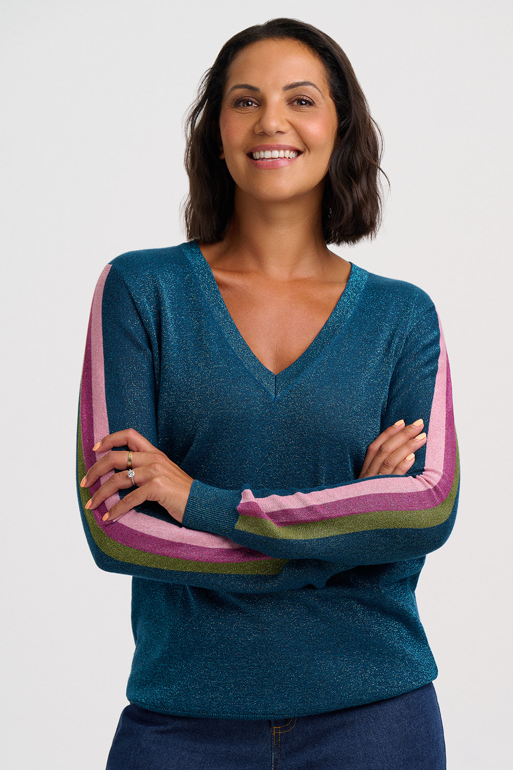 A woman with short dark hair smiling, wearing a teal blue V-neck sweater with pink and olive green striped sleeves, posed with her arms crossed against a light gray studio background.