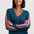 A woman with short dark hair smiling, wearing a teal blue V-neck sweater with pink and olive green striped sleeves, posed with her arms crossed against a light gray studio background.