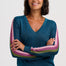 A woman with short dark hair smiling, wearing a teal blue V-neck sweater with pink and olive green striped sleeves, posed with her arms crossed against a light gray studio background.