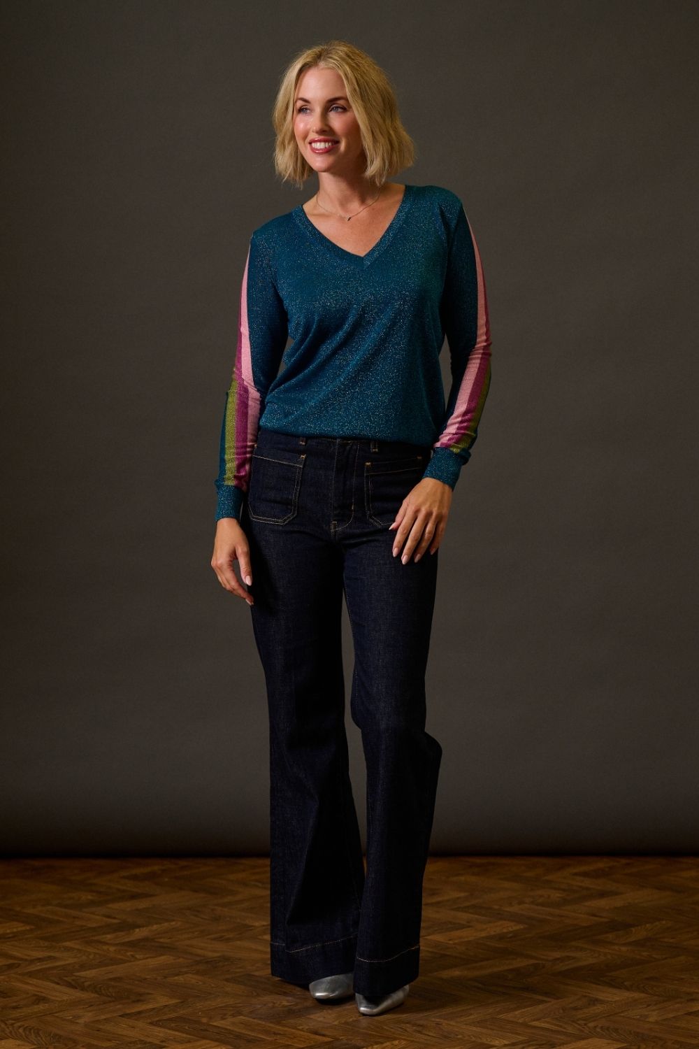 Woman wearing a teal sweater with pink sleeves and dark jeans against a plain background