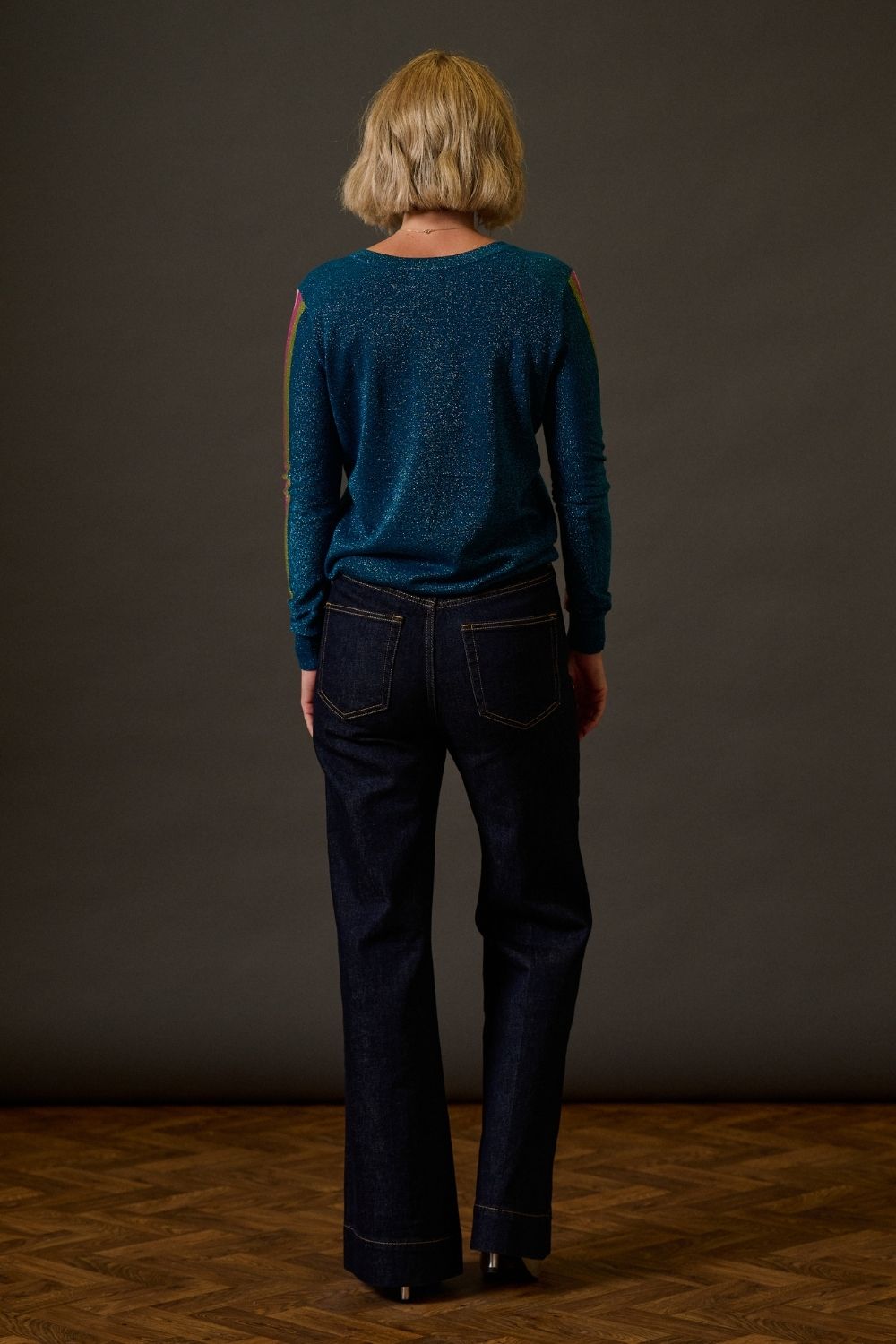 Person wearing a teal sweater and dark jeans standing against a plain background