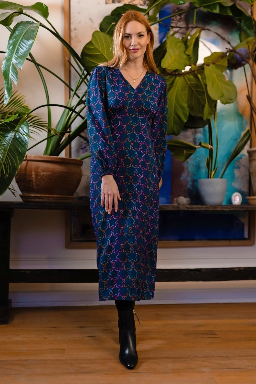 A woman stands indoors wearing a long-sleeve midi dress with a dark blue base and an allover pink and purple geometric pattern. She is wearing black ankle boots, with potted plants and a wooden floor visible in the background.