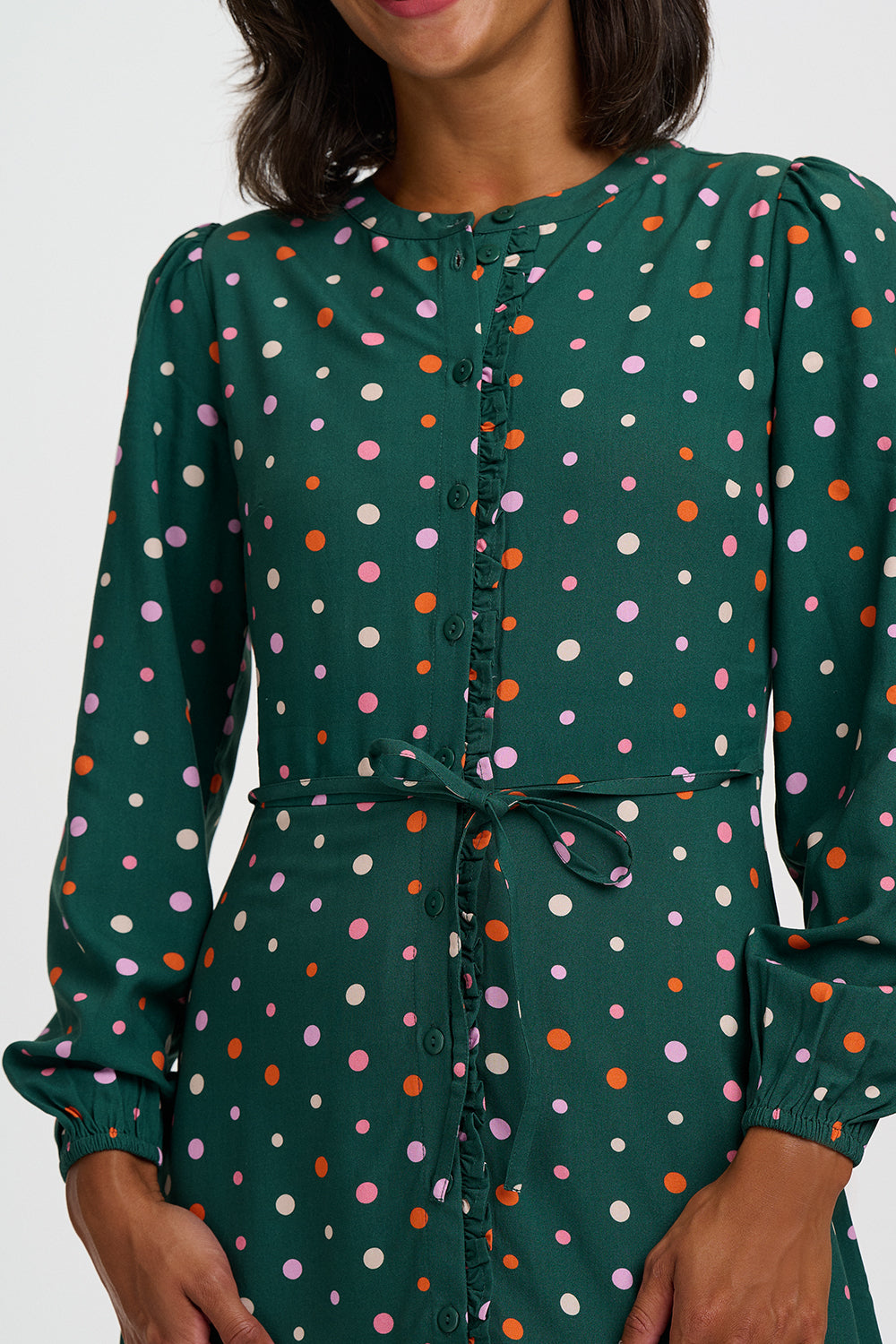A close-up of a woman wearing a dark green long-sleeve dress with a multicolor polka dot pattern and a matching tie belt at the waist.