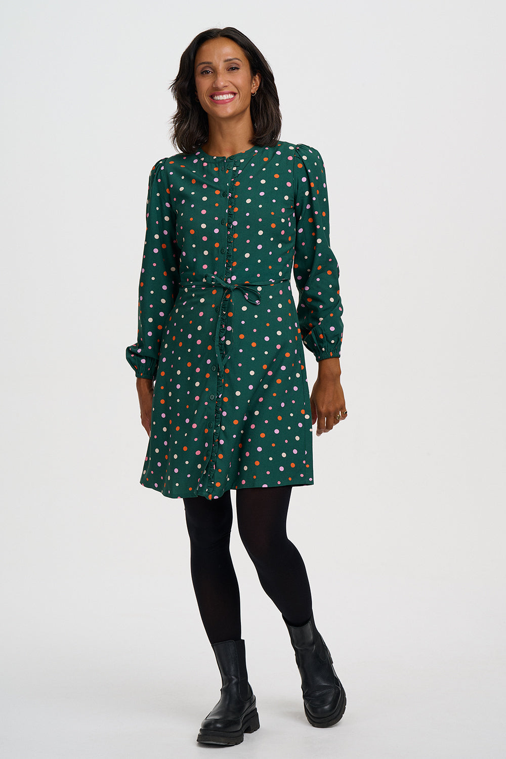 A woman modeling a dark green long-sleeve dress with a white and orange polka dot pattern, paired with black tights and black leather ankle boots, against a plain light gray studio background.