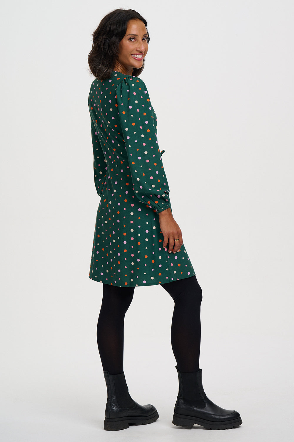 A woman modeling a dark green long-sleeve dress with a colorful polka dot pattern, paired with black tights and black boots, photographed against a plain white studio background.