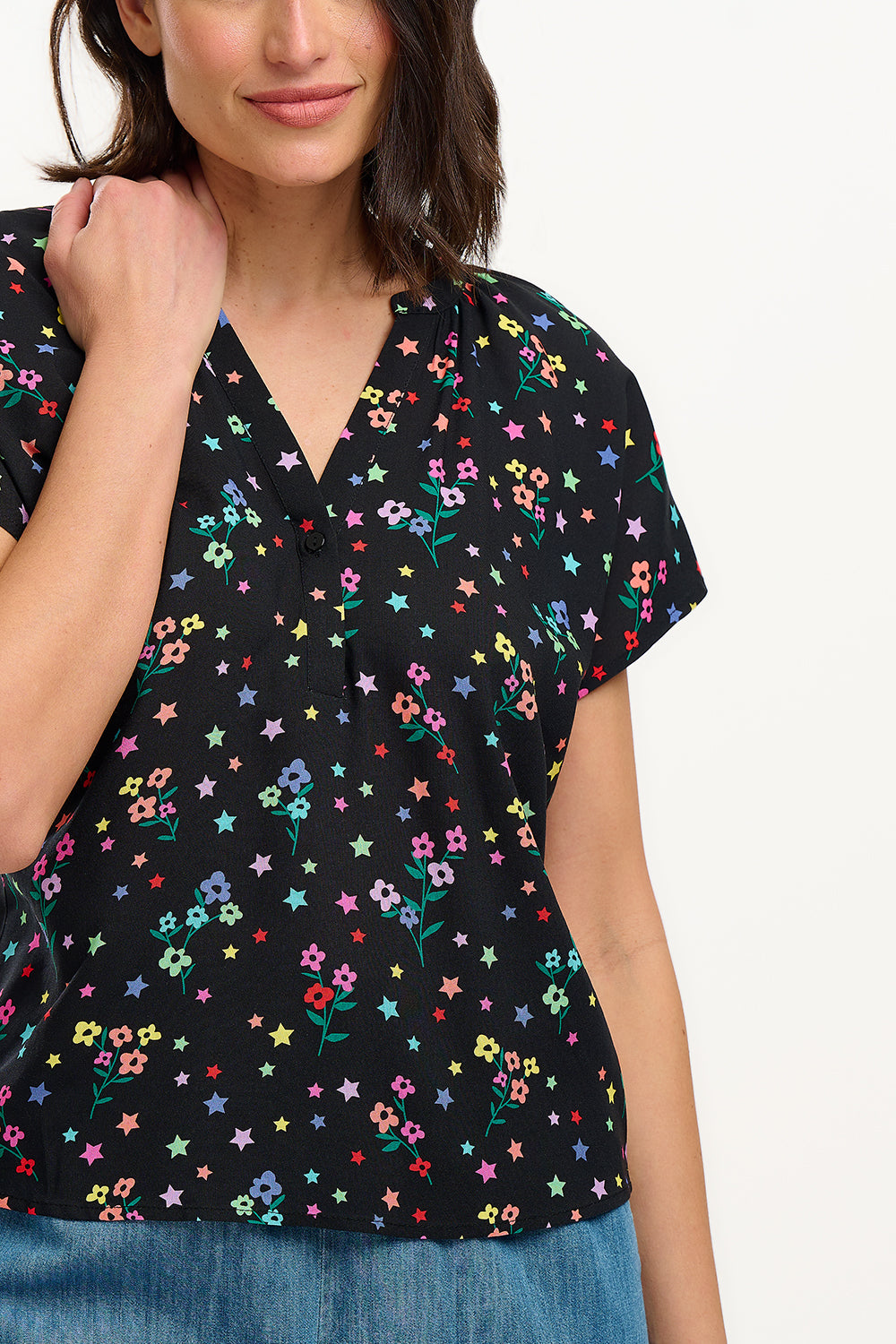A woman smiling and wearing a black blouse with a colorful floral and star print, featuring a round grandad collar and short cap sleeves.