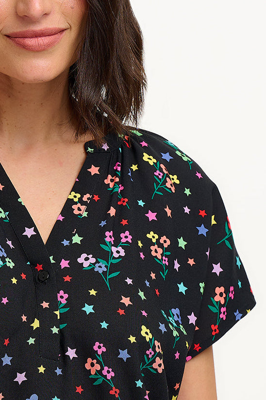 A woman smiling and wearing a black blouse with a colorful floral and star print, featuring a round grandad collar and short cap sleeves.
